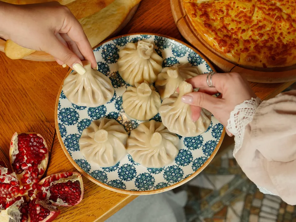 Hands picking up khinkali from a plate with khachapuri and pomegranate nearby